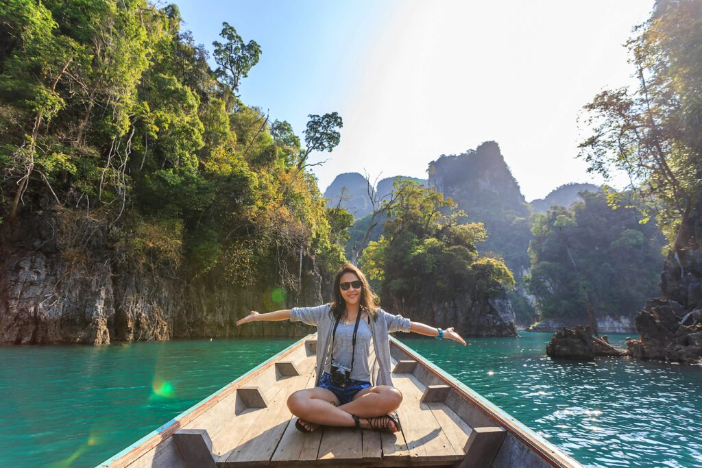A picture of a young lady with her hands stretched out sideways, she's on a boat on the backdrop of a beautiful rainforest, used to depict sustainable tourism.