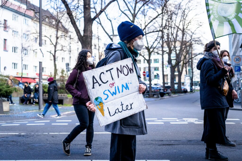 Photo of a young woman in black jacket holding white printer paper written "act now or swim later" to depict COP28 goals