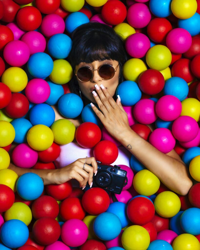 A picture of a lady lying amidst multi colored small round balls, with one hands on her mouth and another holding a camera you may need when preparing for your trip 