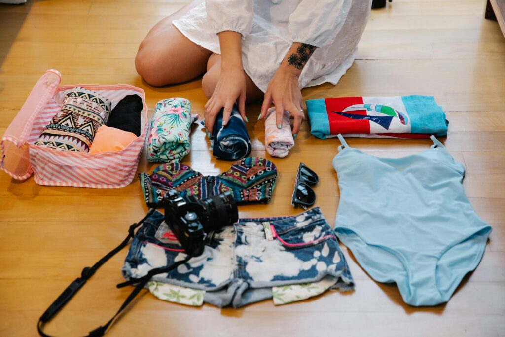 A picture os a lady seated on the floor with her hands holding clothes folded alongside a camera and other travel essentials